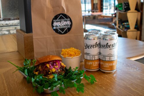 A pulled pork sandwich with coleslaw, mac and cheese, and two cans of beer next to a takeout bag on a wooden table.