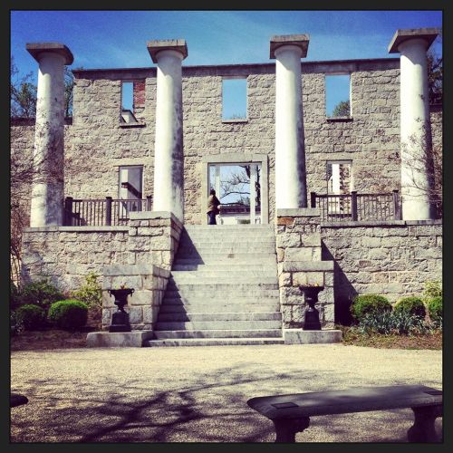 Ruins of a stone building with tall columns, steps leading up, and a person standing in the doorway under a clear blue sky.