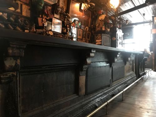 A long, dark wooden bar with ornate details, surrounded by vintage decor and framed photos in a cozy setting.