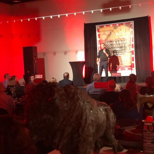 A comedian performs on stage in a red-lit venue, with an audience seated at tables and a lion statue in the foreground.