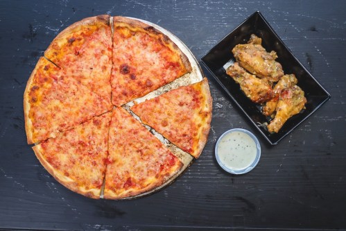 A large cheese pizza with slices cut, next to a plate of chicken wings and a small bowl of dipping sauce.