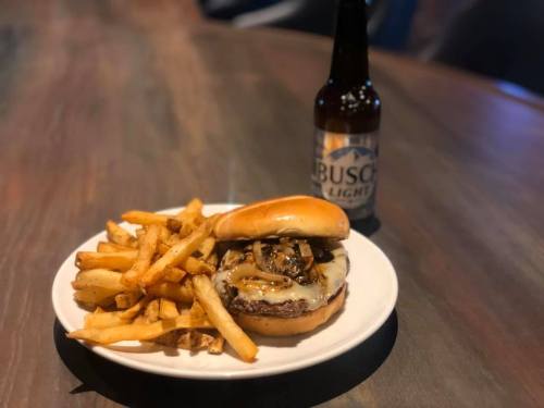 A juicy burger with melted cheese and grilled onions, served with fries and a bottle of Busch Light on a wooden table.