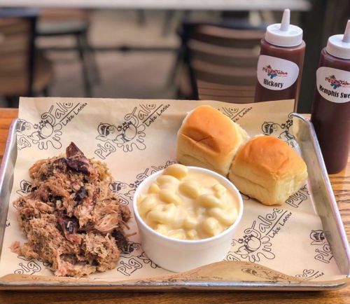 A tray with pulled pork, macaroni and cheese, two dinner rolls, and two bottles of barbecue sauce.