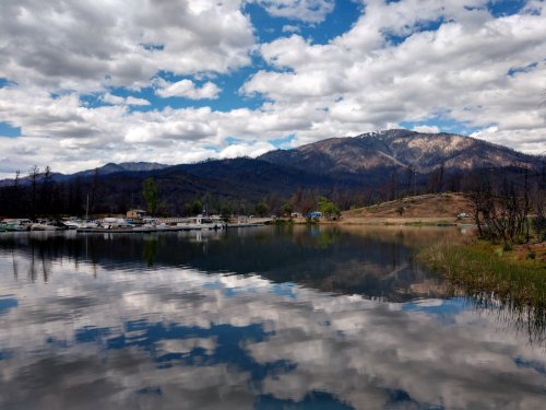 A serene lake reflects clouds and mountains, with boats docked along the shore and charred trees in the background.