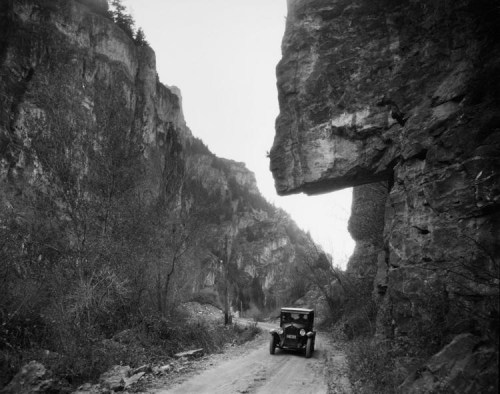 A vintage car drives along a narrow dirt road between towering rock formations in a canyon.