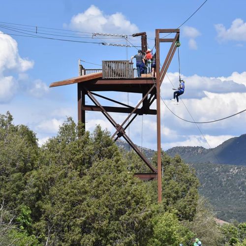 A zip line platform with people preparing to ride, set against a backdrop of mountains and blue skies.