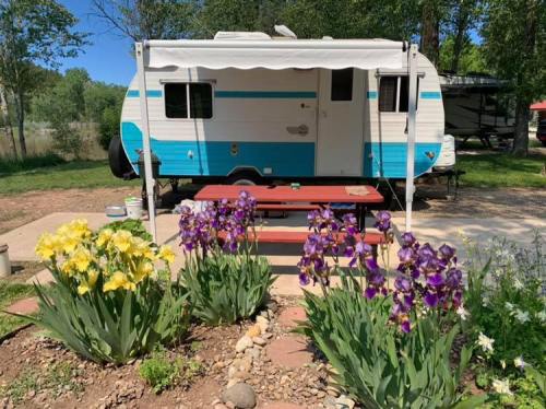 A vintage camper parked near a picnic table, surrounded by colorful flowers and greenery in a sunny outdoor setting.