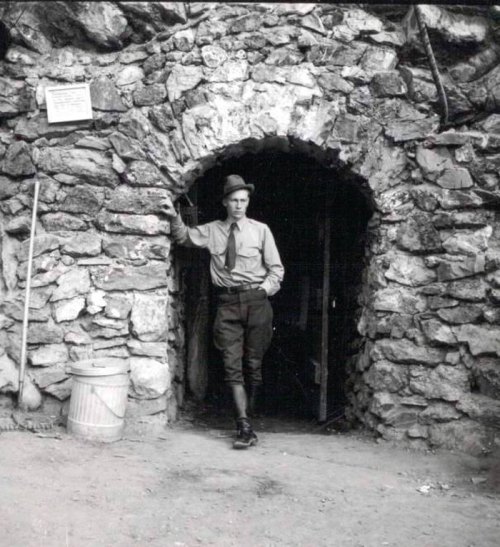 A man in a hat and uniform stands in an archway of a stone structure, with tools and a bucket nearby.