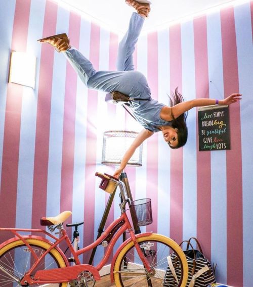 A woman hangs upside down from the ceiling in a colorful room with a pink bicycle and motivational wall art.