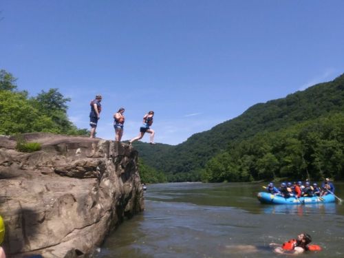 Three people prepare to jump off a rock into a river, while a group in a raft floats nearby surrounded by green hills.