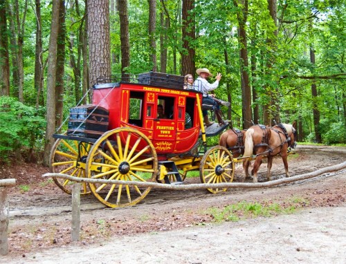 A colorful horse-drawn stagecoach travels through a wooded area, with a driver and passenger waving.