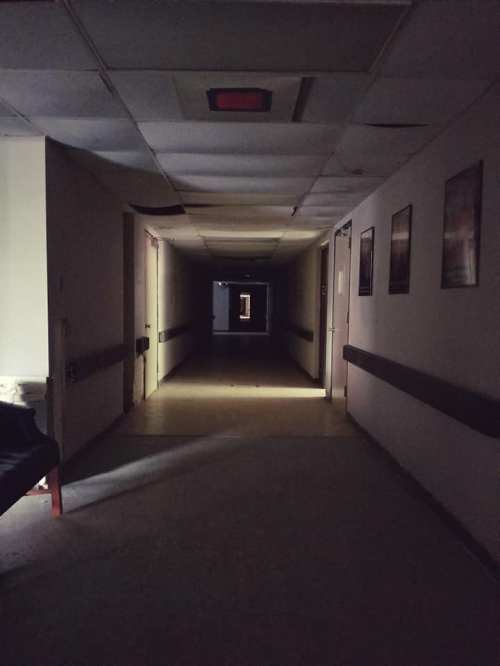 Dimly lit hallway with doors on either side, leading to a darkened area at the end. Shadows create an eerie atmosphere.