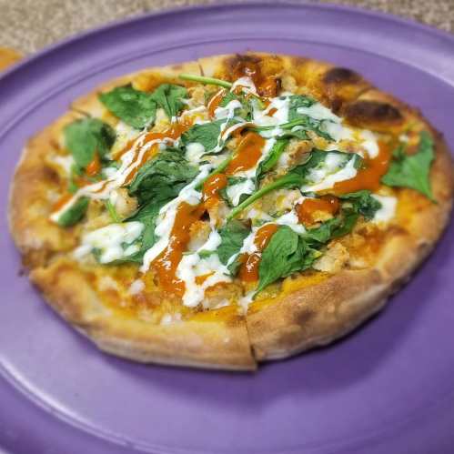 A freshly baked pizza topped with spinach, chicken, and drizzled with sauce on a purple plate.