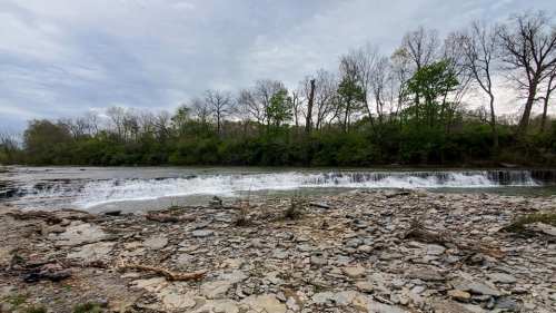 A serene river scene with a gentle waterfall, surrounded by trees and rocky terrain under a cloudy sky.