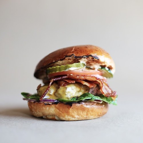 A delicious burger with bacon, lettuce, pickles, and cheese on a toasted bun, set against a neutral background.