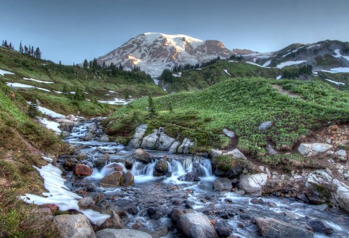 A serene mountain landscape with a snow-capped peak, flowing stream, and lush green meadows.