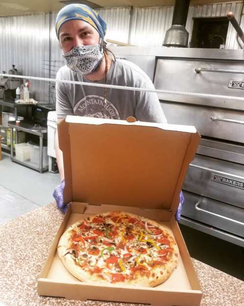 A smiling person in a mask holds a pizza box open, revealing a freshly made pizza with various toppings.