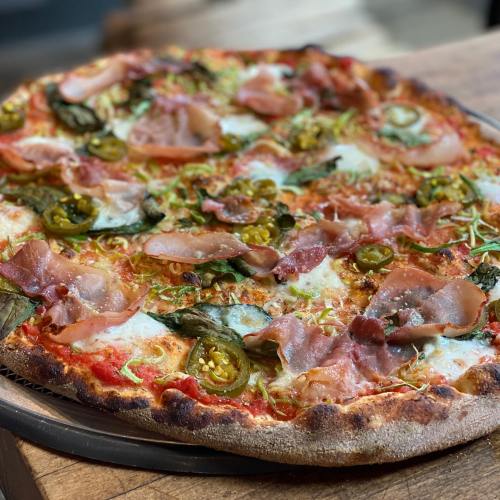 A close-up of a freshly baked pizza topped with ham, jalapeños, and greens on a wooden surface.
