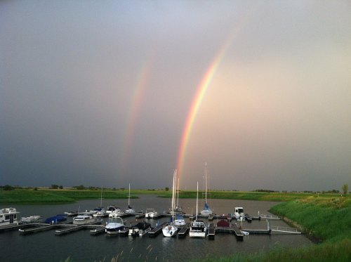 A serene marina with boats, under a sky featuring a double rainbow after a rainstorm. Lush green surroundings.