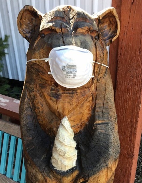 A wooden bear sculpture wearing a face mask, standing outdoors near a colorful bench.