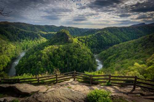 A scenic view of a lush green valley with a winding river, framed by a wooden fence under a cloudy sky.