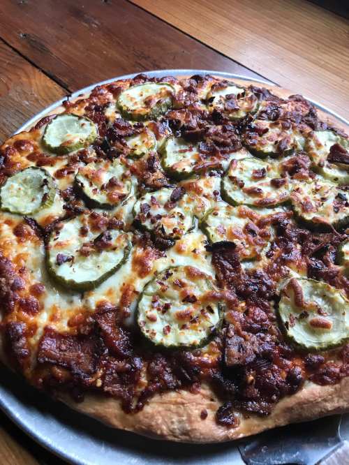 A close-up of a pizza topped with pickles, bacon, and melted cheese on a wooden table.
