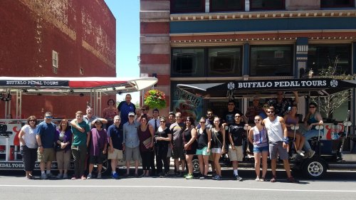 A large group of people poses in front of two pedal tour vehicles on a sunny day in an urban setting.