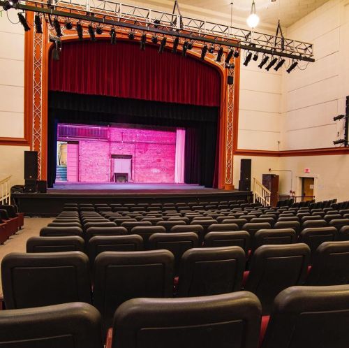 A view of an empty theater stage with red curtains and rows of black seats in the foreground.