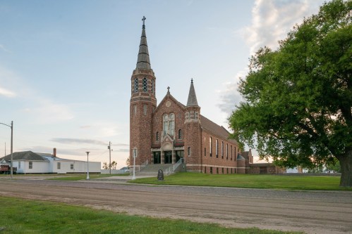 A brick church with a tall steeple stands beside a grassy area and dirt road under a clear sky.