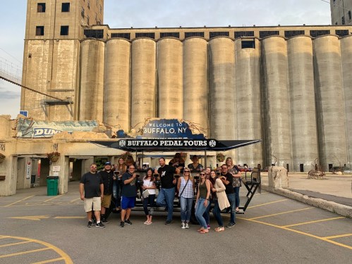 A group of people poses in front of a pedal tour vehicle in Buffalo, NY, with grain silos in the background.