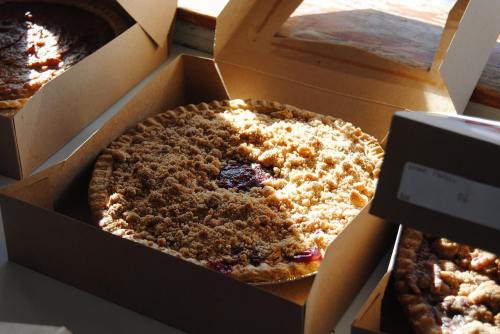 A freshly baked pie with a crumbly topping, placed in a brown cardboard box, illuminated by natural light.