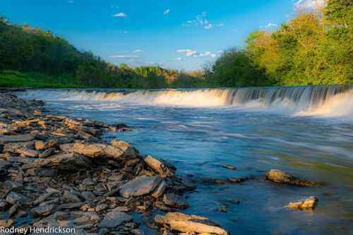 A serene river flows over rocks, surrounded by lush greenery and a clear blue sky with scattered clouds.