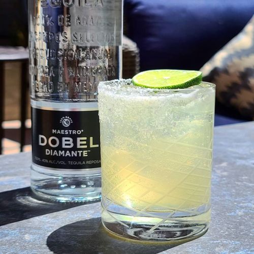 A refreshing cocktail with a lime wedge, served in a glass beside a bottle of Maestro Dobel Diamante tequila.