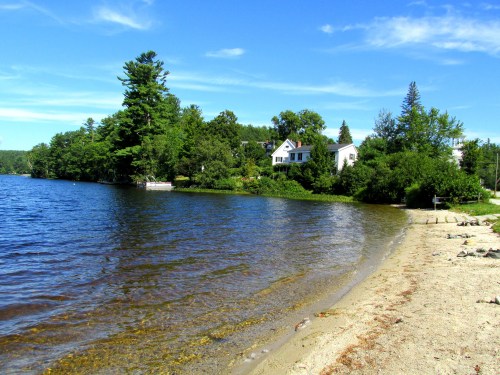 A serene lakeside scene with a sandy shore, lush greenery, and a white house under a clear blue sky.