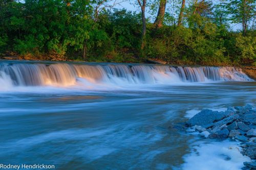 A serene river scene with a gentle waterfall, surrounded by lush greenery and soft blue water.