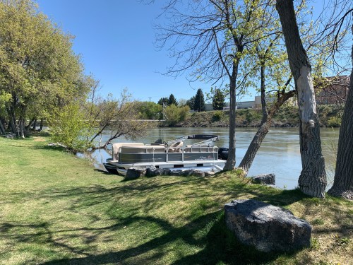 A pontoon boat is docked by a river, surrounded by green grass and trees under a clear blue sky.