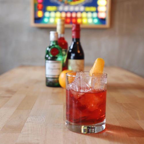 A cocktail glass with ice and an orange twist, with bottles of spirits blurred in the background on a wooden table.