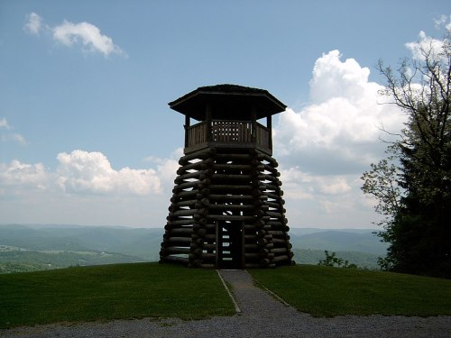 A wooden observation tower on a grassy hill, surrounded by rolling hills and a cloudy sky.