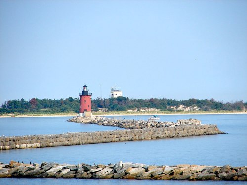 A red lighthouse stands on a rocky jetty, with calm waters and a distant shore in the background under a clear blue sky.