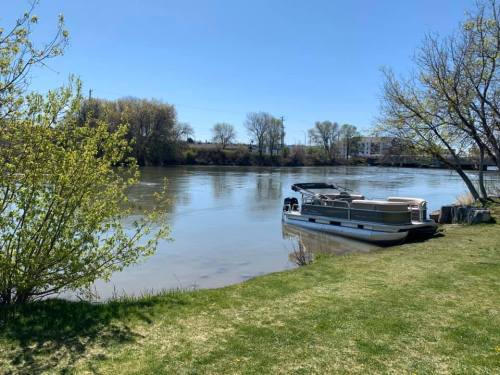 A calm river scene with a boat docked on the shore, surrounded by green grass and trees under a clear blue sky.