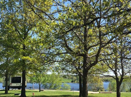 A serene lakeside view with green trees, a picnic area, and an RV parked nearby under a clear blue sky.
