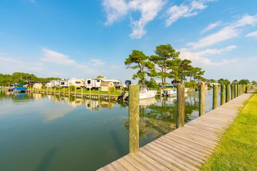 A serene marina with boats and RVs along a calm waterway, surrounded by green trees and a clear blue sky.