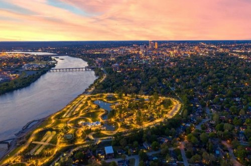 Aerial view of a city at dusk, featuring a river, park with lights, and a skyline in the background.