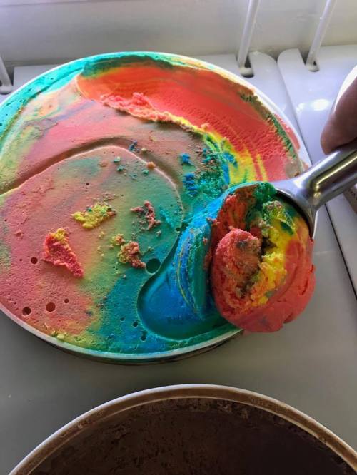 A colorful scoop of rainbow ice cream being served from a round container.