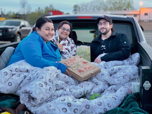 Three friends sitting in the back of a truck, wrapped in blankets, enjoying pizza together in a parking lot.