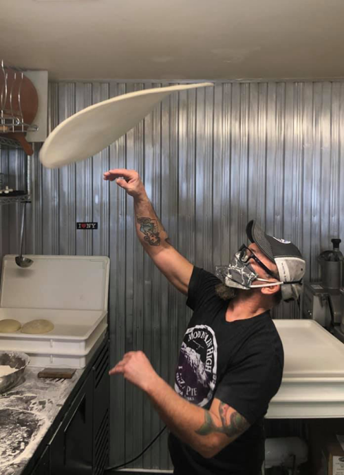 A chef in a mask tosses pizza dough in a kitchen, showcasing skill and focus.
