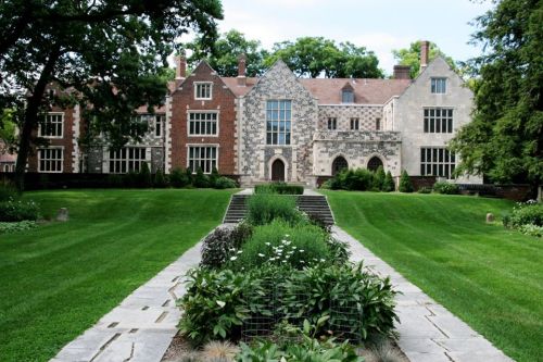 A large, historic mansion with a stone facade, surrounded by lush green lawns and neatly arranged flower beds.