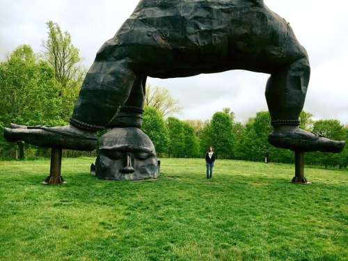A person stands beneath a large, abstract sculpture of a giant figure with oversized feet in a green park.