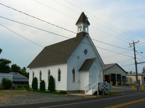 A small white church with a steeple, surrounded by greenery, located near a road with power lines in the background.