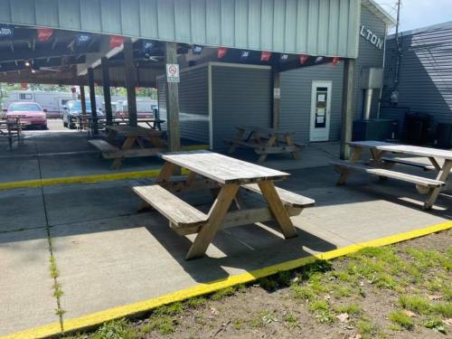 Outdoor seating area with wooden picnic tables under a covered structure, surrounded by a paved walkway and grassy area.